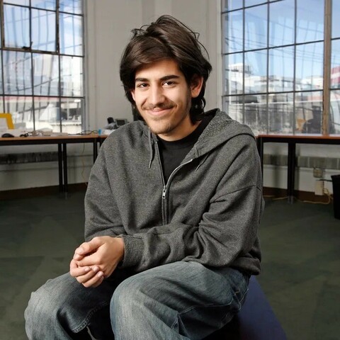 Aaron Swartz sitting on a stool in a loft, smiling (cute), large windows behind him, through which blurred blue sky and blurred buildings are seen 