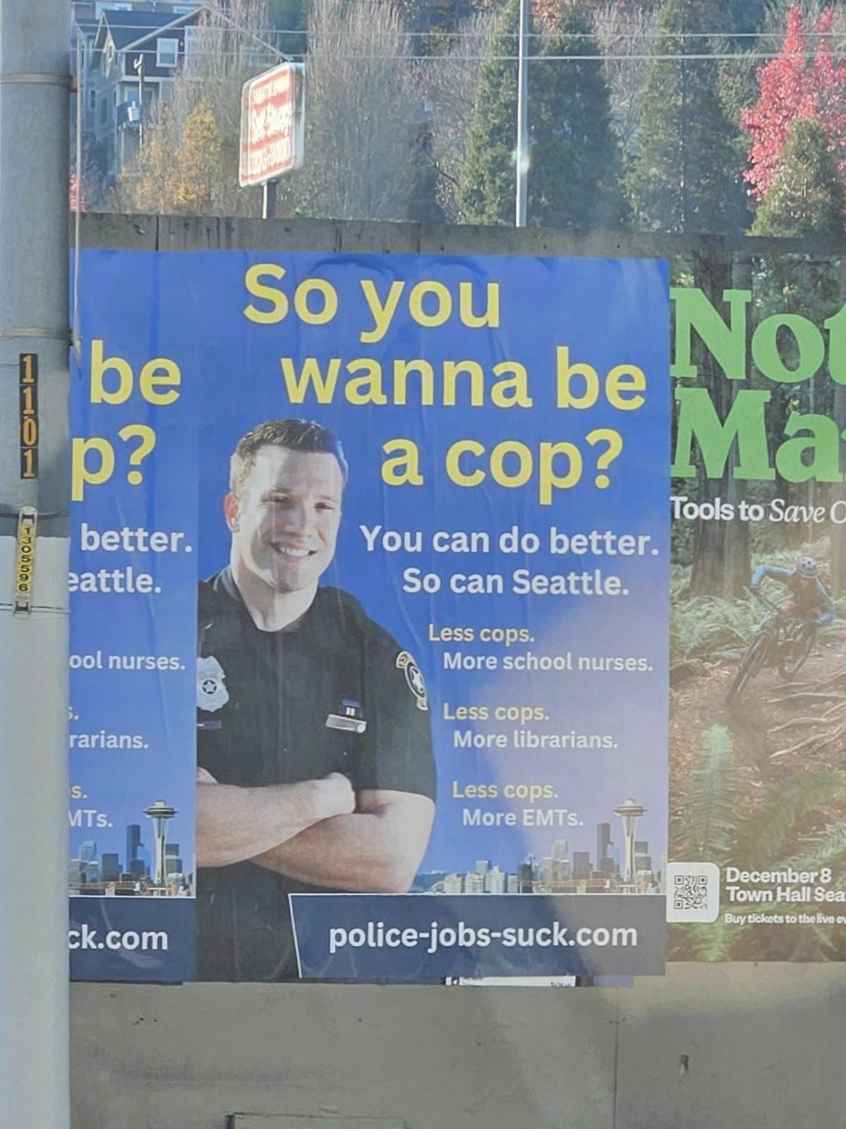 Picture of a poster with a smiling white cop with his arms folded. The Seattle skyline is in the background. The text reads "So you wanna be a cop? You can do better. So can Seattle. Less cops. More school nurses. Less cops. More librarians. Less cops. <br />More EMTs. police-jobs-suck.com"