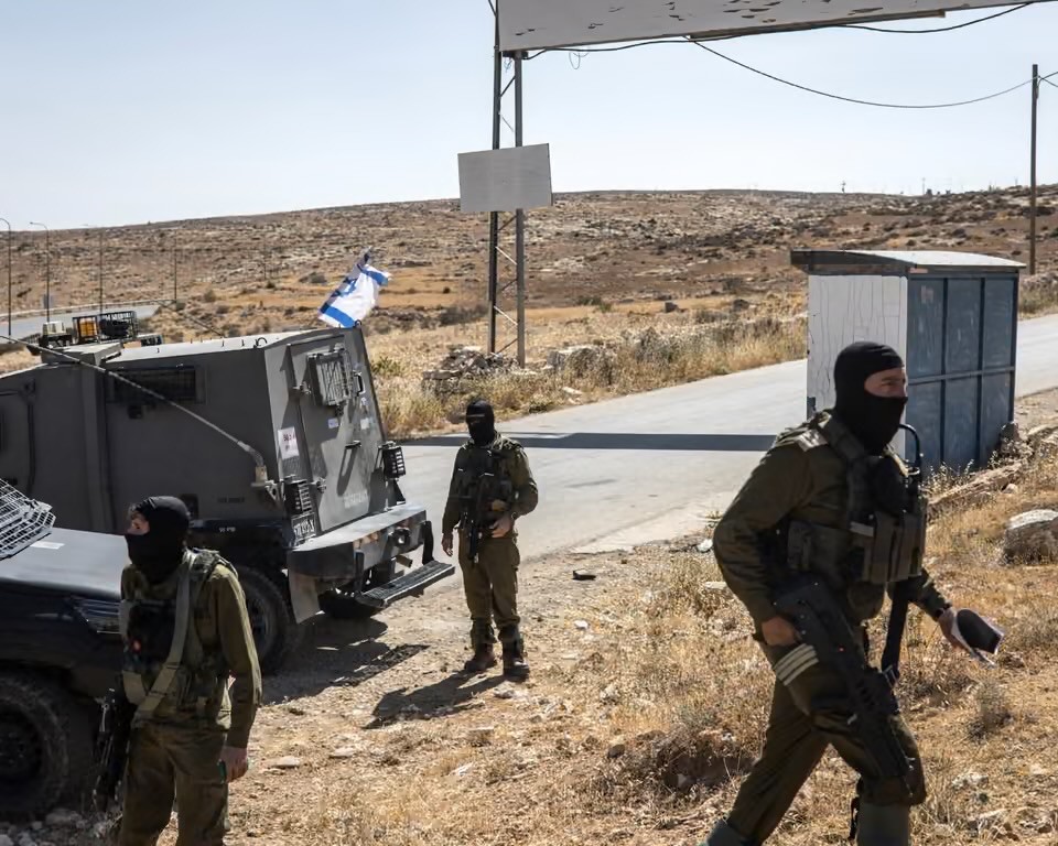 A group of armed soldiers, wearing uniforms and face masks, stands near a military vehicle on a dirt road. An Israeli flag is visible on the vehicle. The background features a dry, hilly landscape and a small shelter.