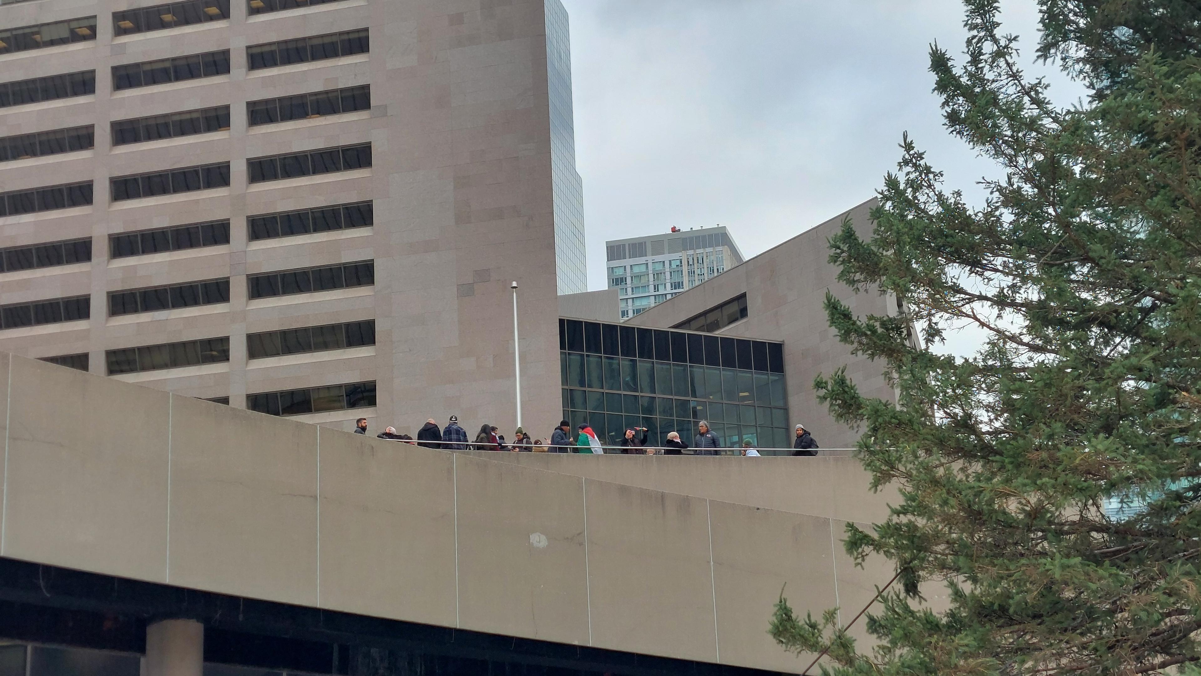 A partial view of a modern building complex (Toronto City Hall) is seen against a cloudy sky. The building features a mix of straight and angled architectural lines and is predominantly beige in color. A group of people are standing along the edge of a flat roof section of the building, appearing to look out over a distant view. Large, dark-tinted windows are visible along the angled portions of the structure. A pine tree branch occupies the right side of the frame, partially obscuring the view.
Provided by @altbot, generated privately and locally using Gemma3:27b
🌱 Energy used: 0.420 Wh