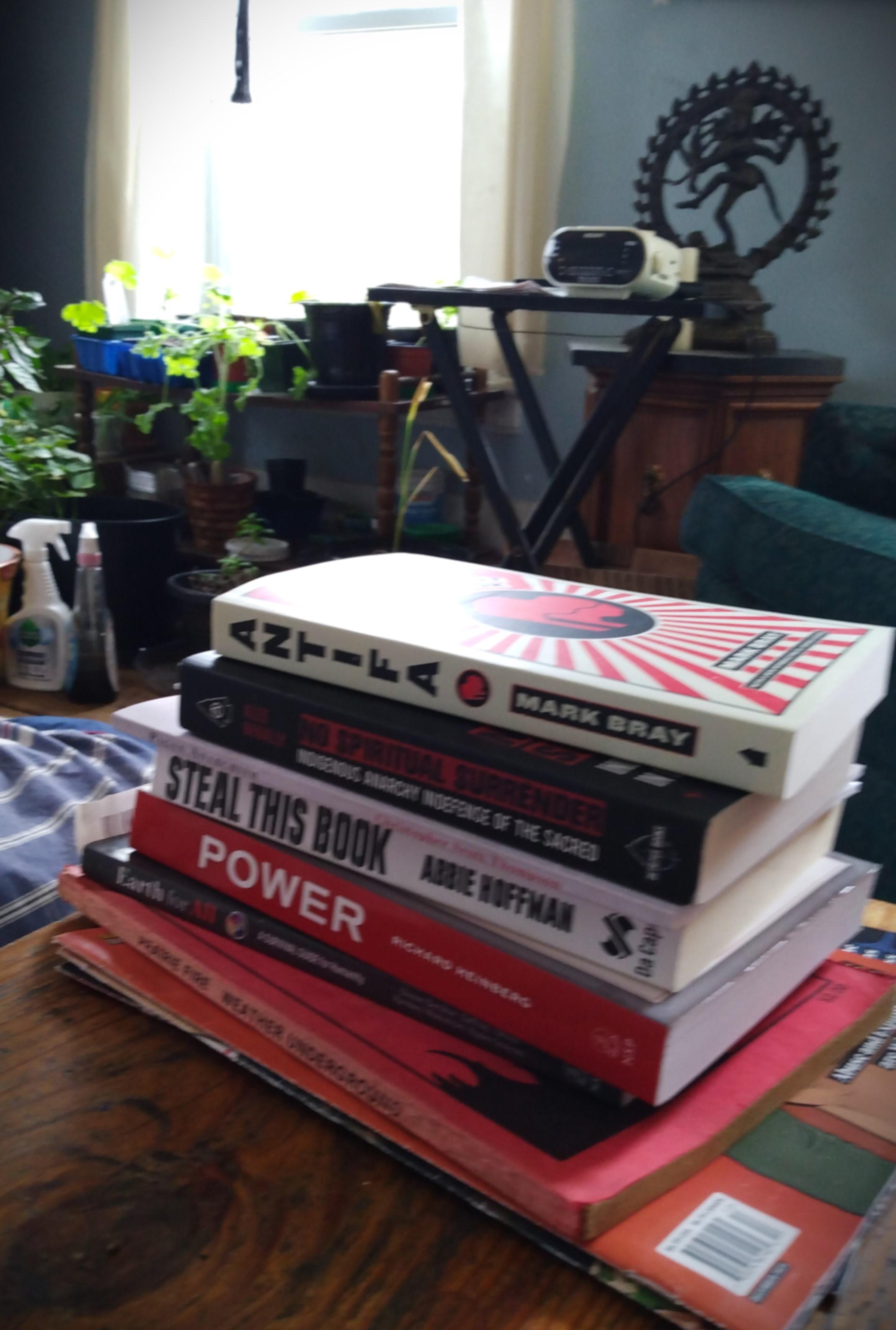 A pile of books on a wood coffee table. In the background is a folding table, a table with plants, and a pedestal with a statue of the Hindu god Shiva on top.

Book titles:
Antifa - The Anti-Fascist Handbook by Mark Bray
No Spiritual Surrender - Indigenous Anarchy in Defense of the Sacred by Klee Benally
Pagan Anarchism by Christopher Scott Thompson
Steal This Book by Abby Hoffman
Power by Richard Heinberg
Earth For All - A Survival Guide For Humanity, A Report to the Club of Rome by Sandrine Dixson-Dedeleve, et al.
Prairie Fire by The Weather Underground
In These Times (Socialist 'zines)