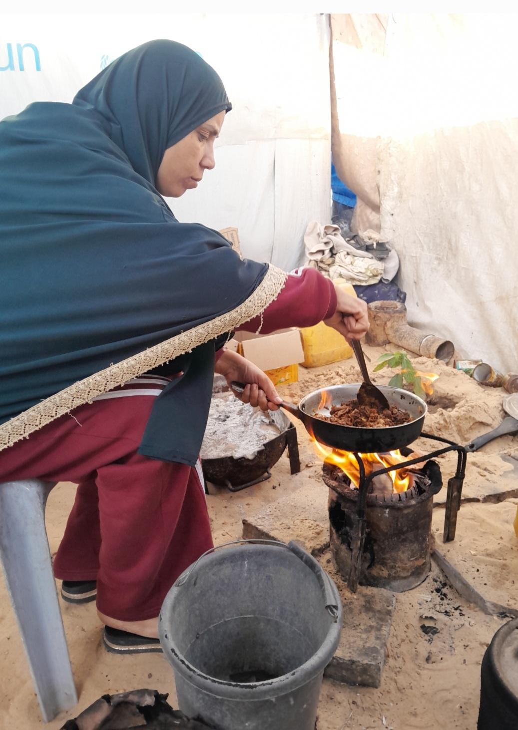 A woman wearing a gray hijab sits in a chair, stirring food in a pan over a fire.