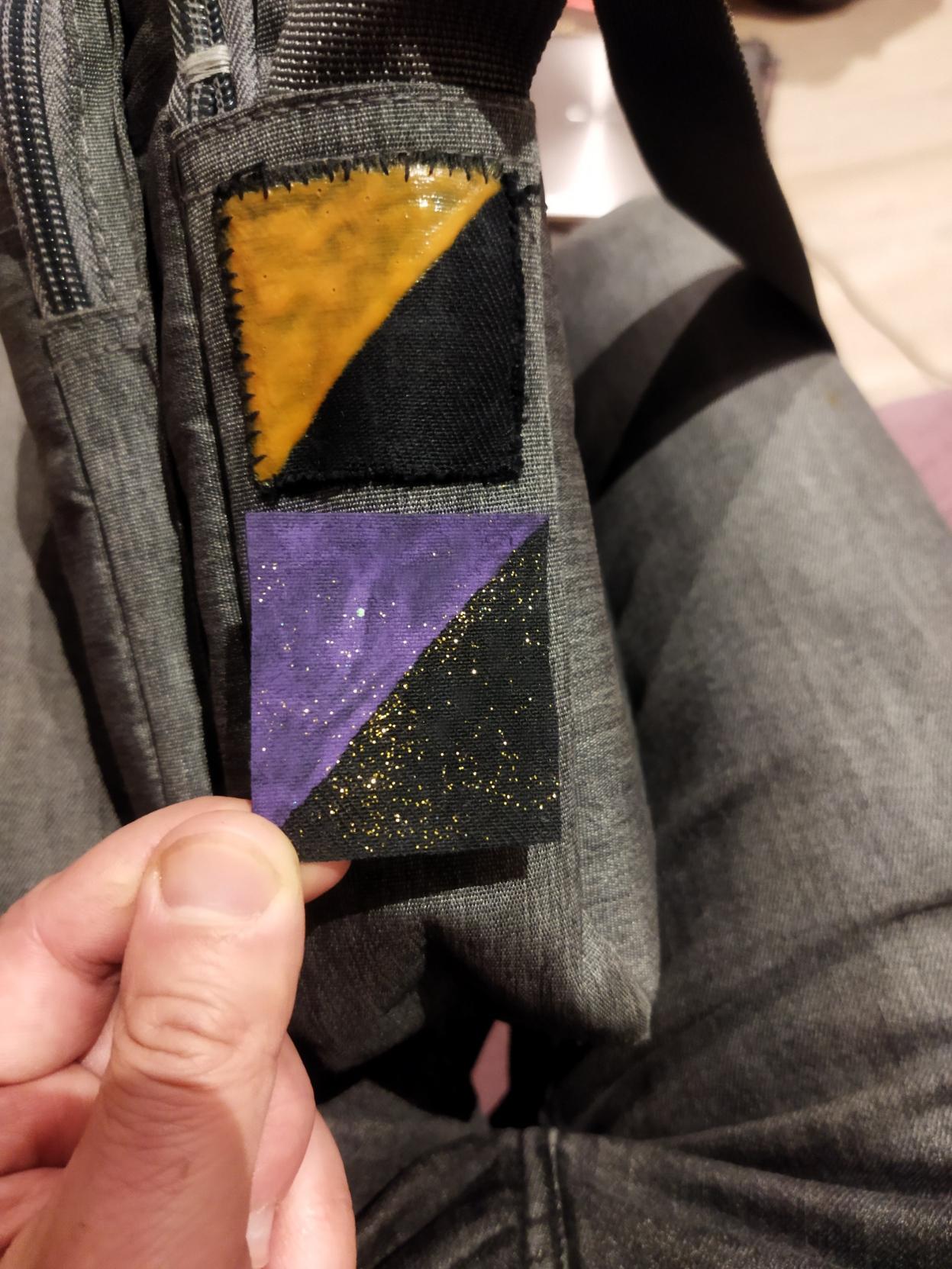 A gray bag side with orange-black patch sawn on and a purple-black patch fitted held with fingers.