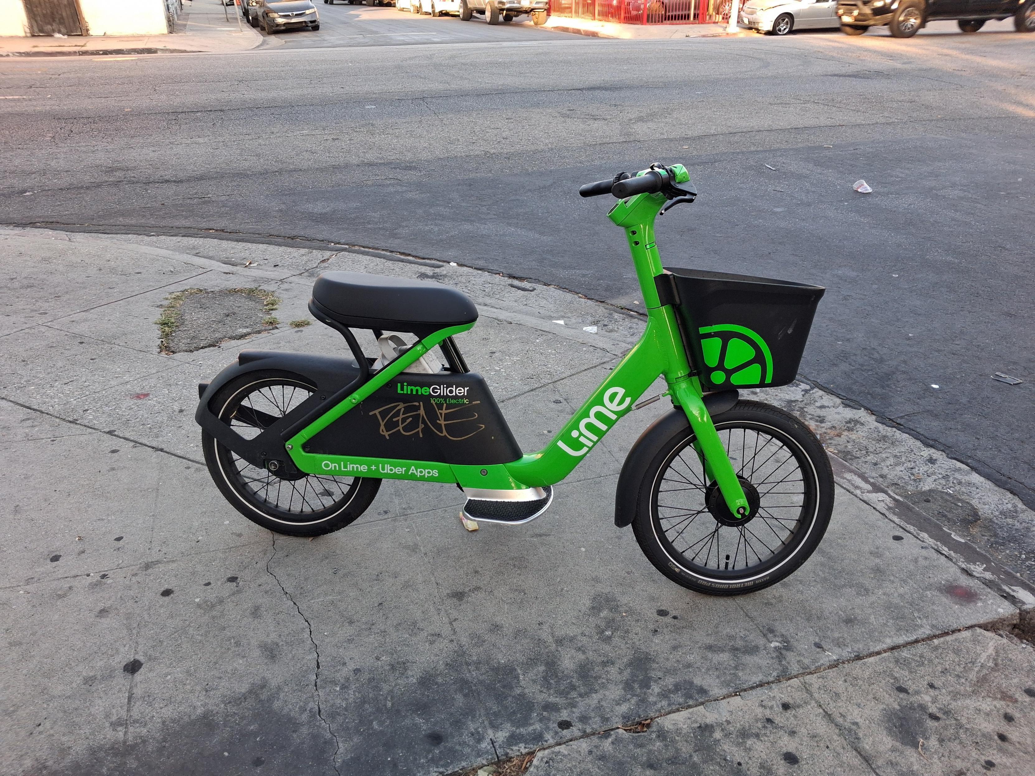 A side view of a green Lime electric bike parked on a street corner. There is a curb cut behind it and a lot of black grease stains of exactly the sort left by an illegal pupusa stand, which is exactly what they are, in fact.