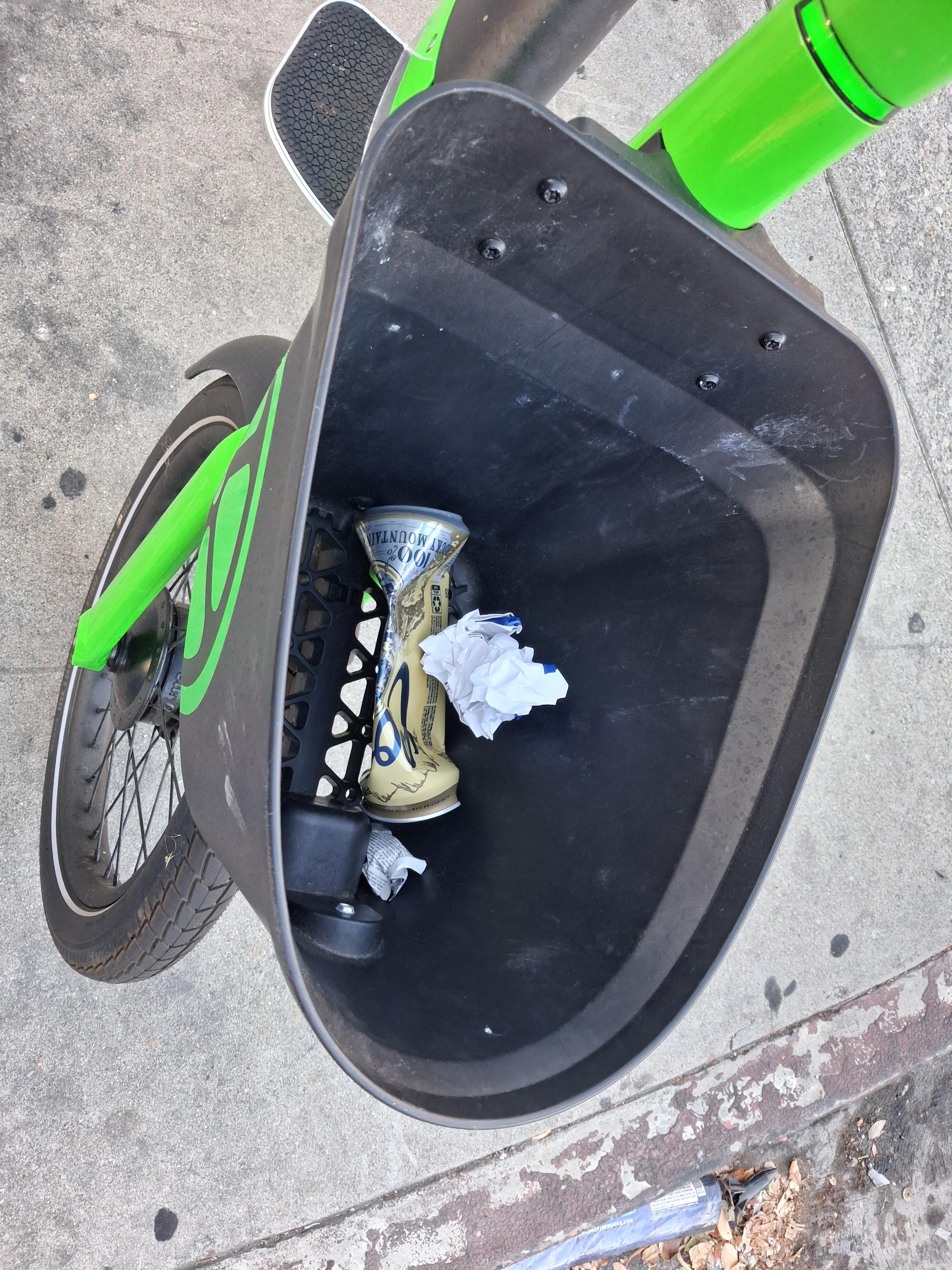 A downward view into the bike's handlebar basket showing an assortment of litter, including a semismooshed and presumably empty Coors can.