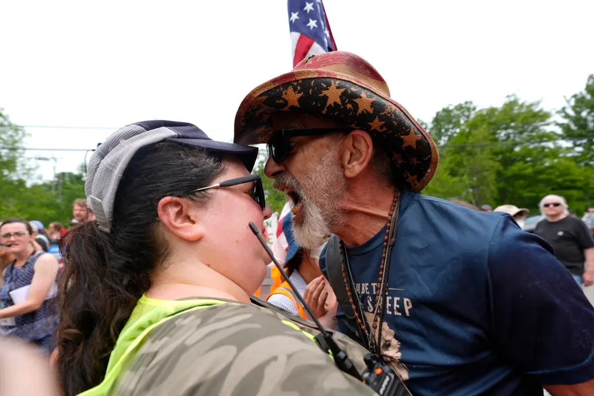A close up of two people at a rally. One is a dark-haired woman holding a walkie talkie and wearing a lime-green vest. The other is an angry white man wearing a "patriotic" cowboy hat. The man is yelling at the woman, showing off his yellow teeth. The man looks like he's about to bust a vein in his red neck.