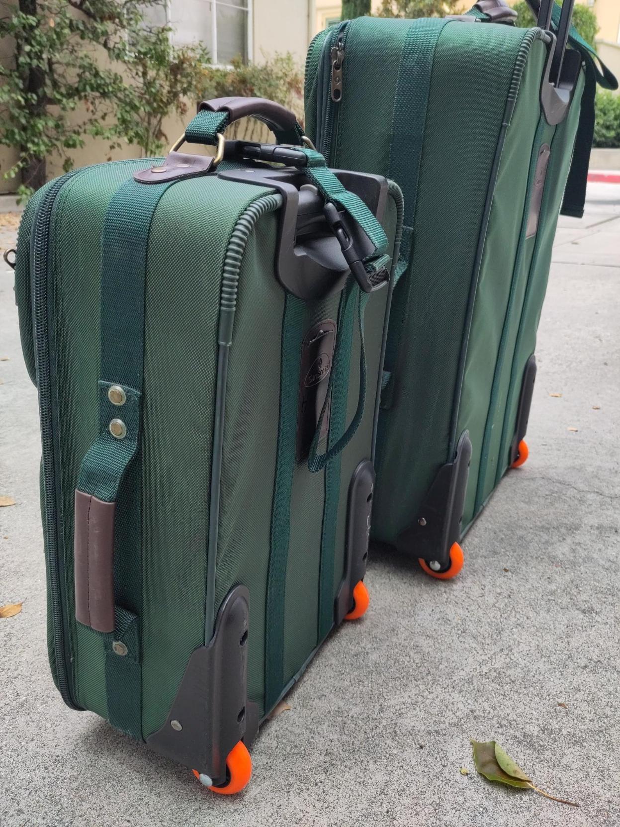 A pair of emerald-green roller bags, one smaller than the other, facing away from the camera so as to show off their new hot orange wheels. Pictured outdoors in a paved area.
