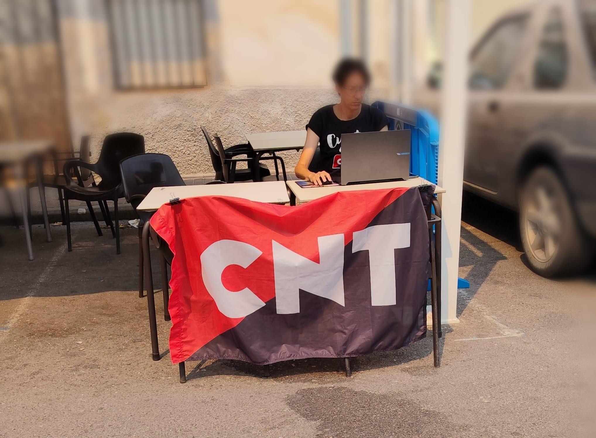 Fotografía de una mesa improvisada, con la bandera rojinegra de CNT y un compañero sentado con su portátil. De fondo se ven más mesas y sillas.