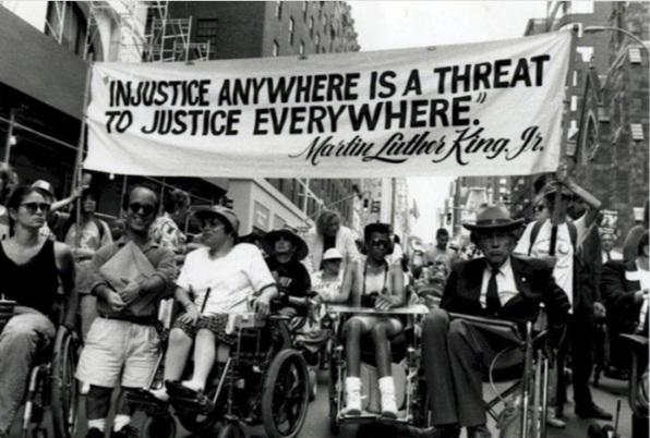 People in wheel chairs in the street demonstrating for passage of Americans With Disabilities Act beneath a banner with the following quote by MLK: Injustice Anywhere is a Threat to Justice Everywhere.
