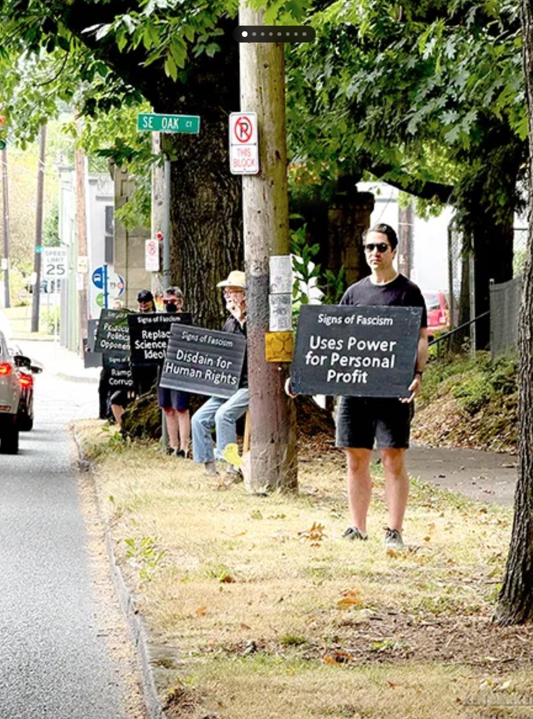Photo of people standing along a street, holding signs with white lettering on black backgrounds, each sign says, "Signs of Fascism" followed by different signs: 
Uses Power for Personal Profit
Disdain for Human Rights
Replaces Science with Ideology (this person is wearing a respirator mask)
Persecutes Political Opponents (this person is wearing a hat that says Fuck ICE, and is holding the sign in front of their face)
Rampant Corruption
Scapegoats Vulnerable Minorities