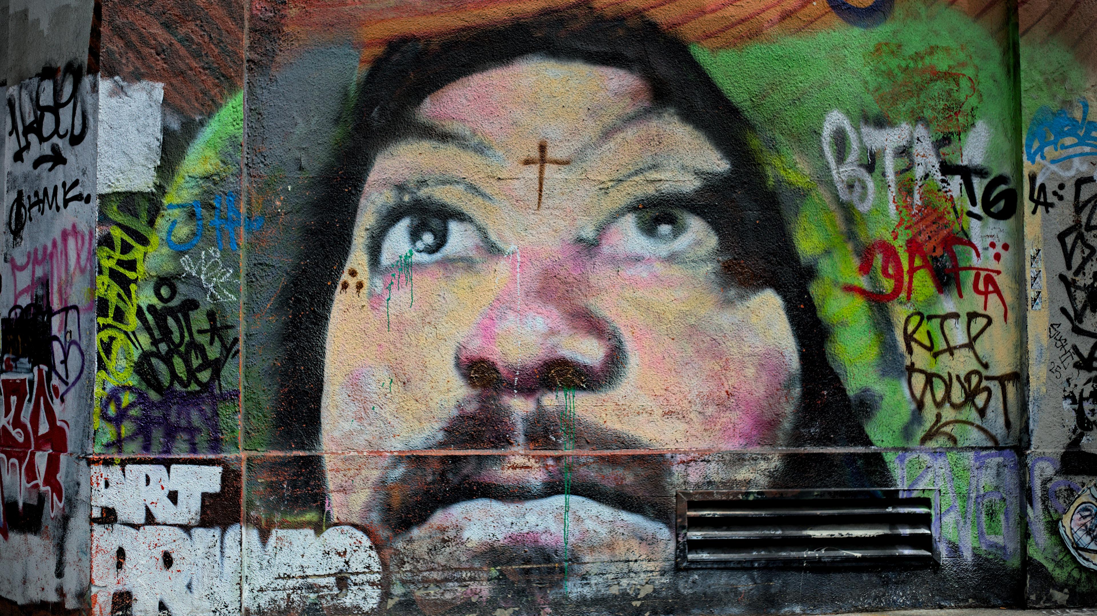 Unknown mural of a person's face at 11th ave and e pine street, male presenting black hair and mustache, looking up, cross in center of forehead, surrounded by multi color graffiti