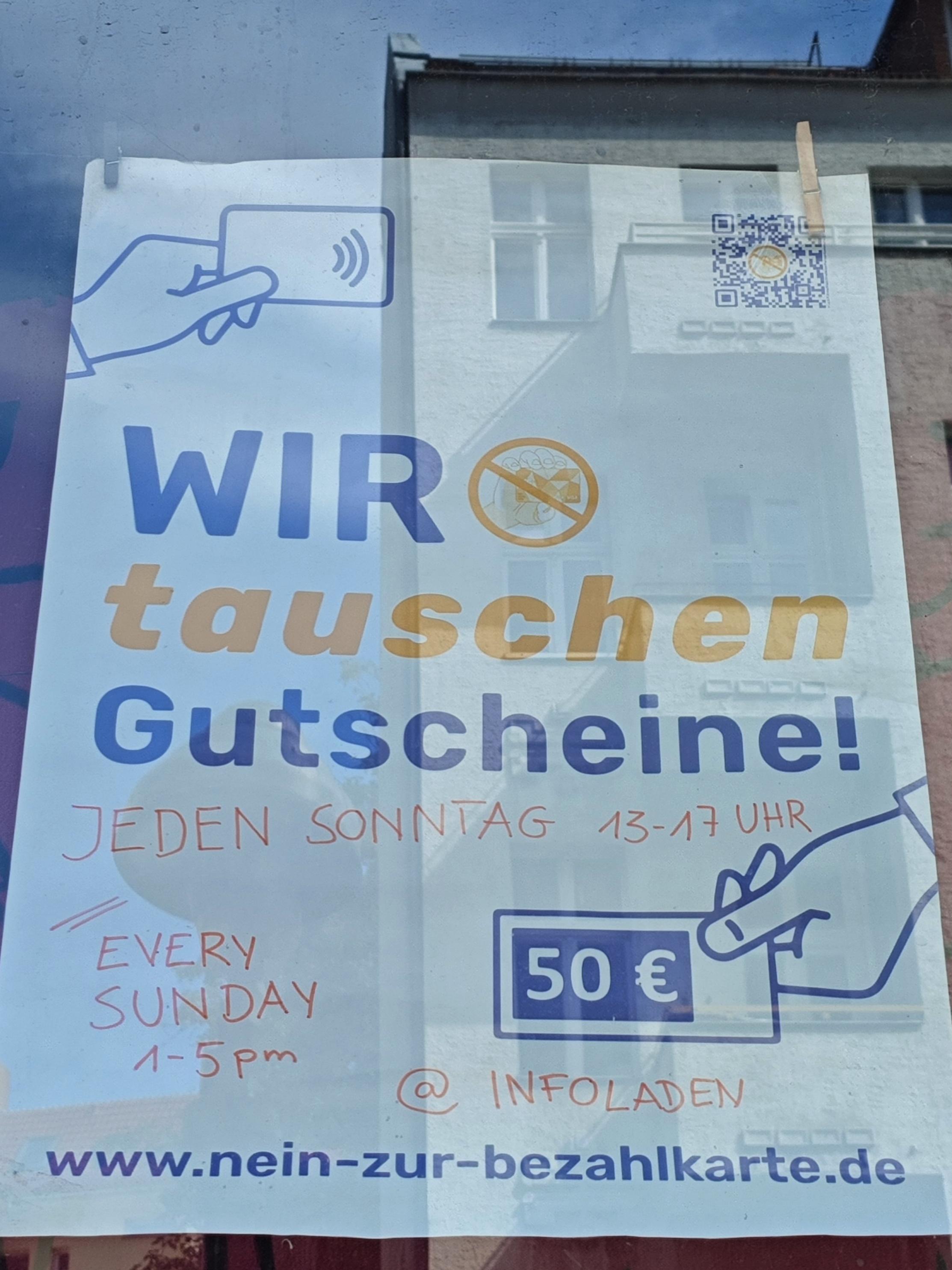 Foto eines Posters zur Bezahlkarte hinter einer Scheibe. Es steht die Zeit zum Umtausch drauf und die Internetadresse. Es ist schematisch eine Hand abgebildet, die einen 50€-Schein hält. Der Text "Wir tauschen Gutscheine".