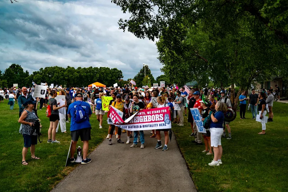 Large protest outside in a park with signs and banners in support of immigrants and against Greg Bovino white nationalist rally.