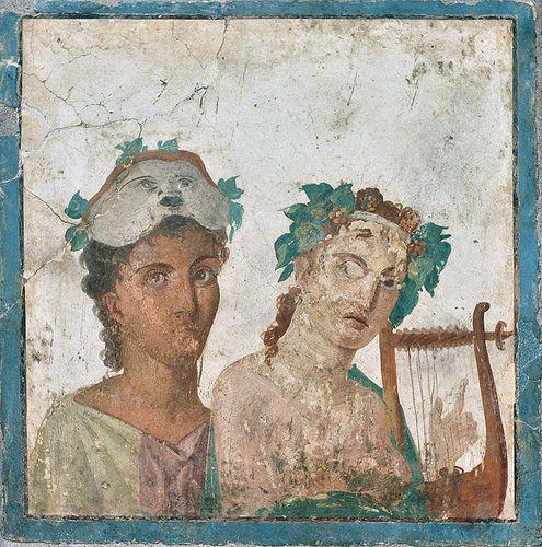 Roman fresco with two figures next to each other. The one to the left has a theatre mask pushed up and resting on their head and looks towards the viewer. The figure to the right holds a lyre and appears to be looking at the figure to the left with an expression of ‘what just happened?’ Fresco from Pompeii and now held by the MAN Napoli.