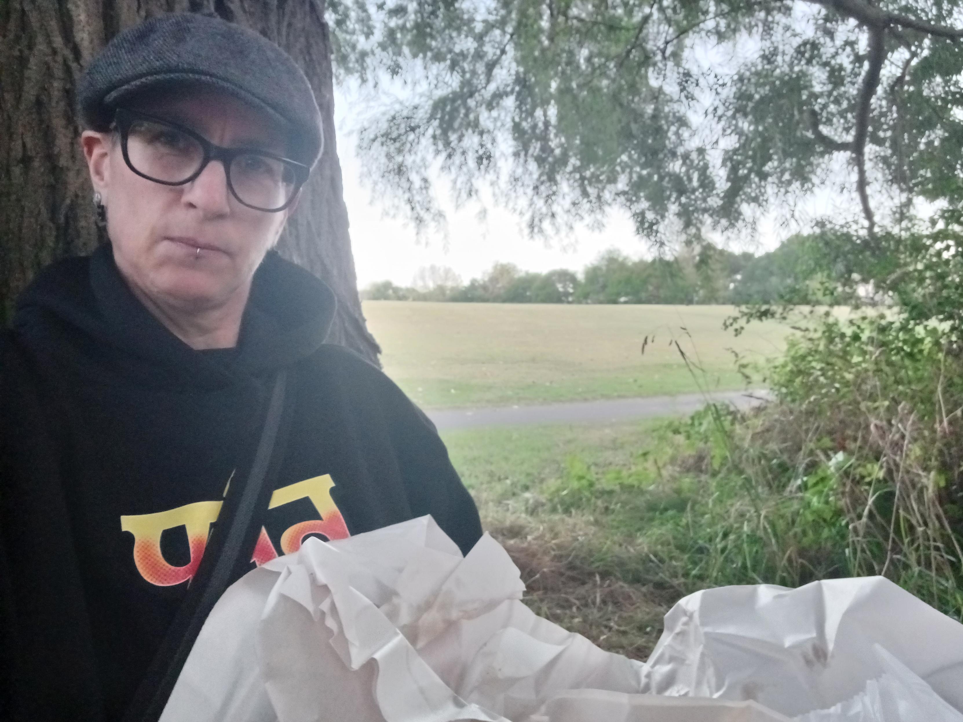 me sitting on my tree seat with food. i am wearing a black 'the mountain' gorillaz hoodie, black rimmed glasses & a north-yorkshire cap. your can see the field behind me.