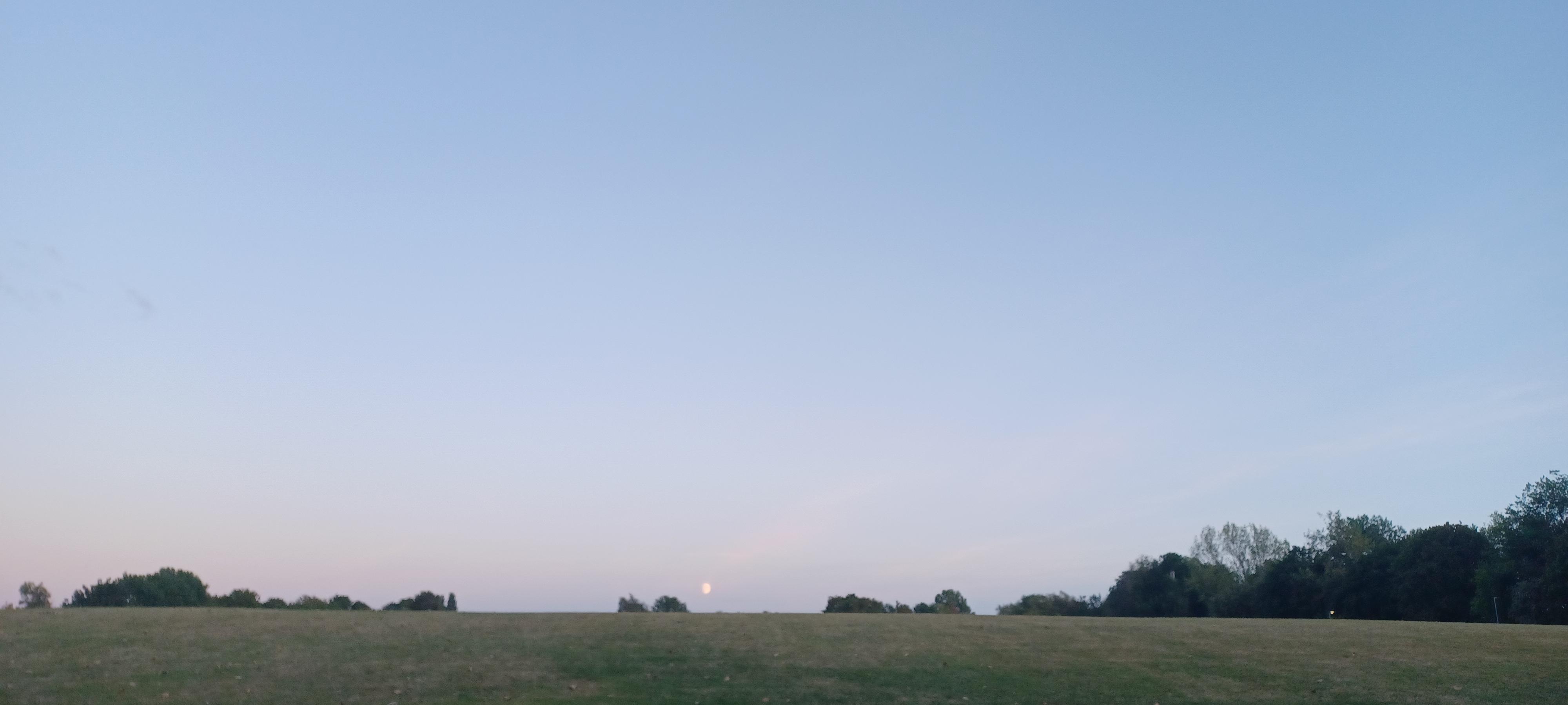 the moon is just visible above the rim of the hill with a few trees just peeping up from behind the hill with a huge blue sky above